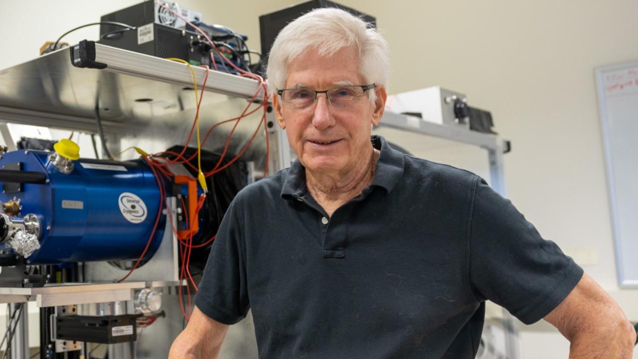 Older man with white hair and glasses smiling in a lab workshop with machinery