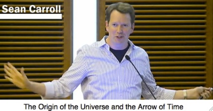 A man gestures while speaking at a lecture, with wooden panels in the background.